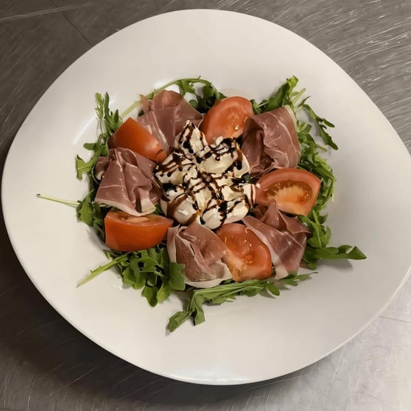 A white plate of Italian salad with prosciutto, tomatoes, burrata cheese, arugula, and balsamic glaze.