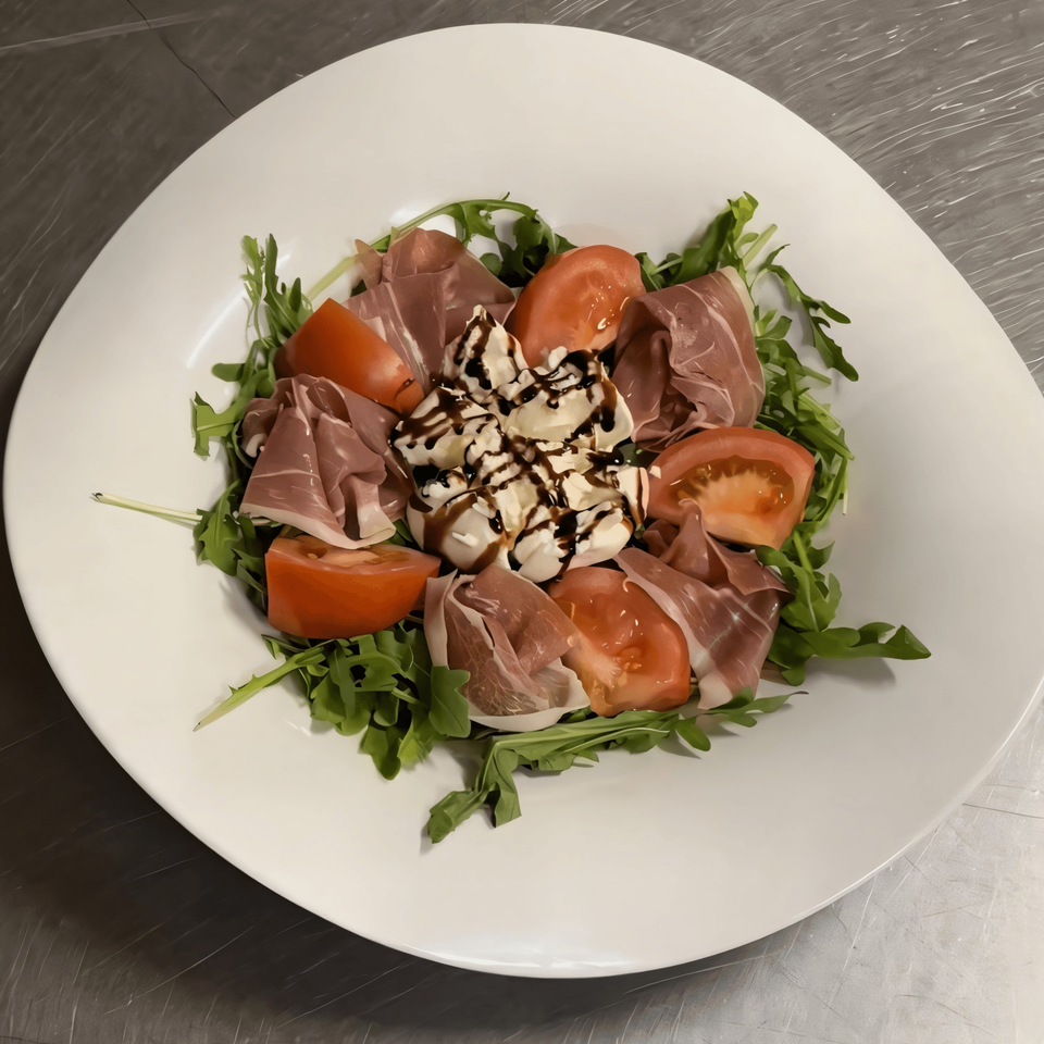 A white plate of Italian salad with prosciutto, tomatoes, burrata cheese, arugula, and balsamic glaze.