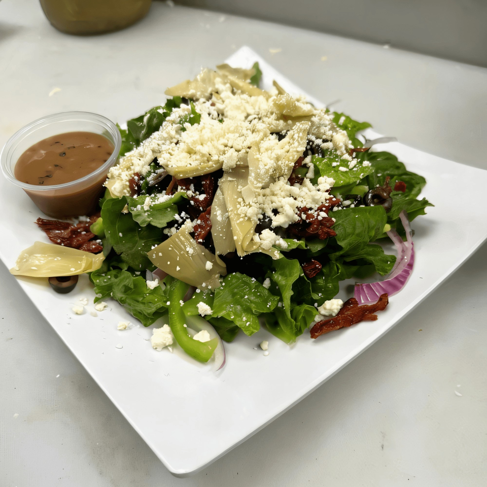 A fresh Italian salad with lettuce, artichokes, sun-dried tomatoes, feta, and dressing on a white plate.