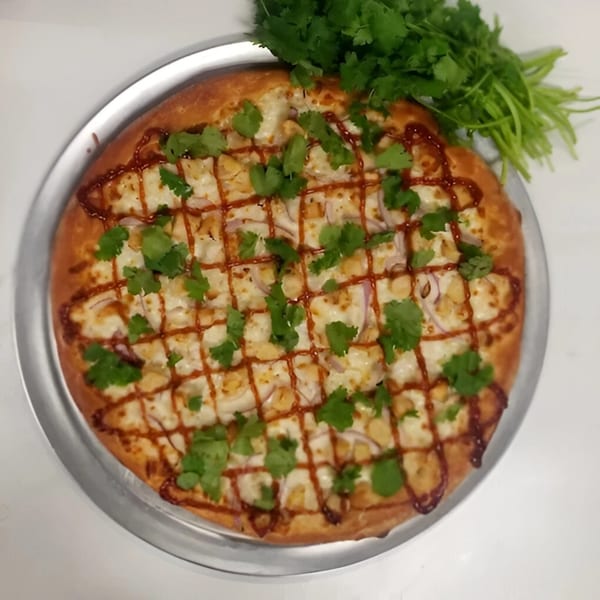 A BBQ chicken pizza with red onions, cilantro, and a drizzle of BBQ sauce on a metal pan.